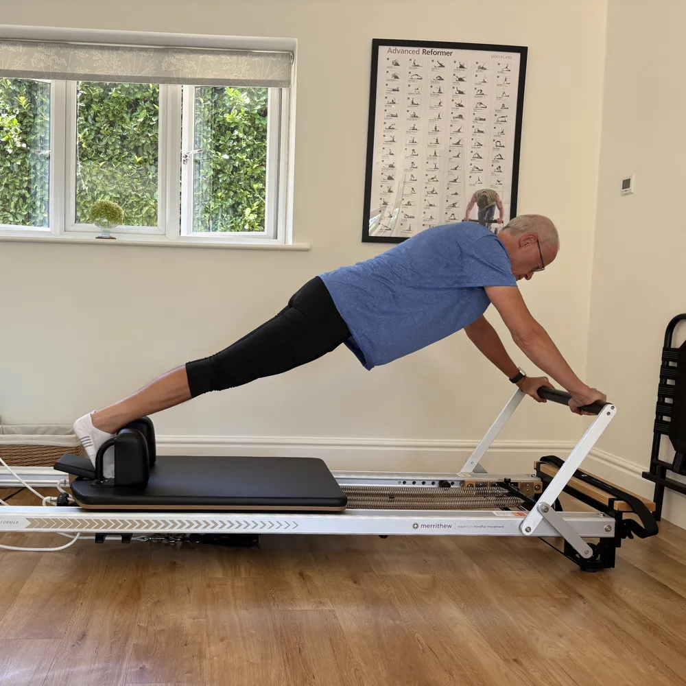 A client in a long plank along the Reformer, hands on the footbar and feet against the shoulder rests