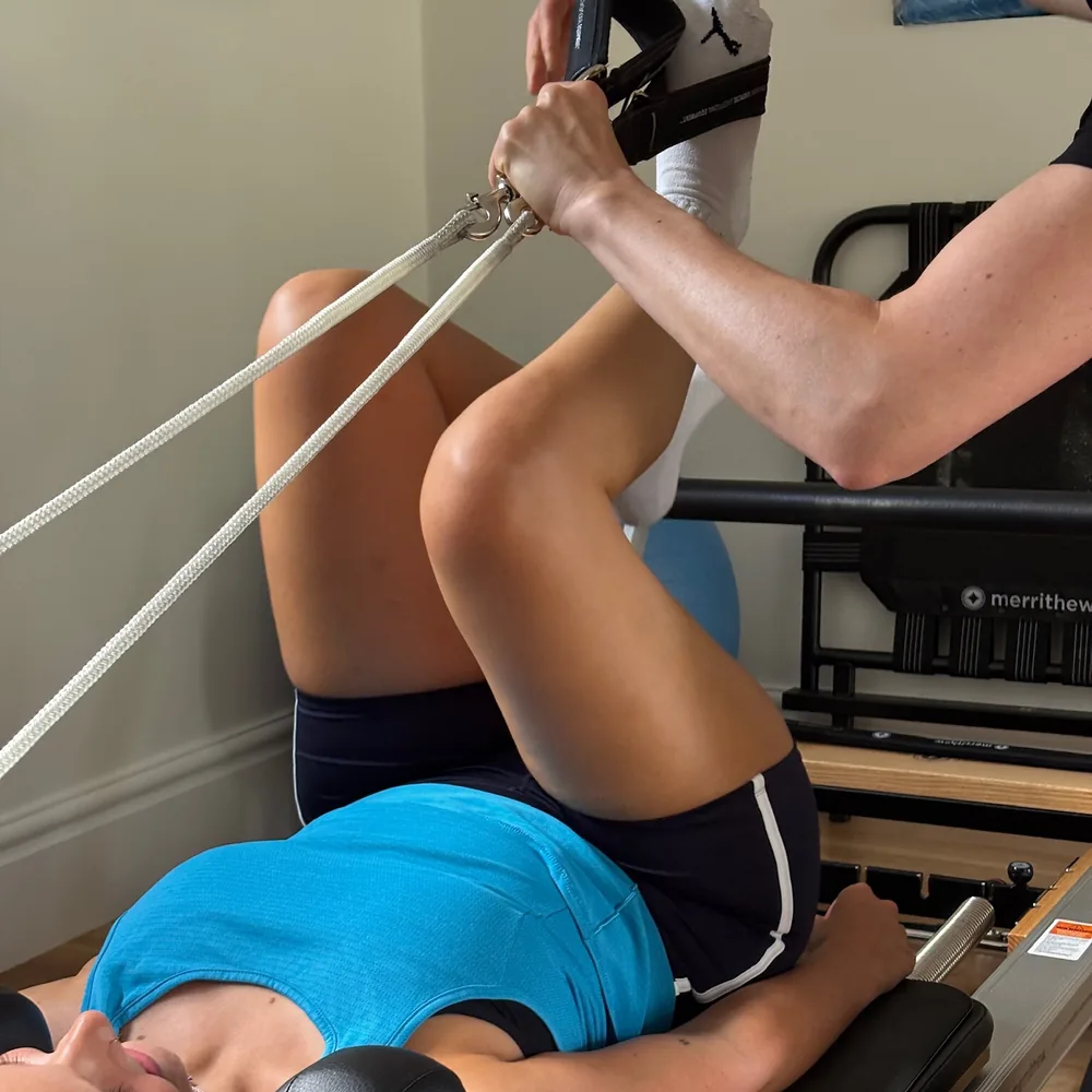 Natasha guiding a client’s foot into the Reformer strap, the ropes running back to the carriage