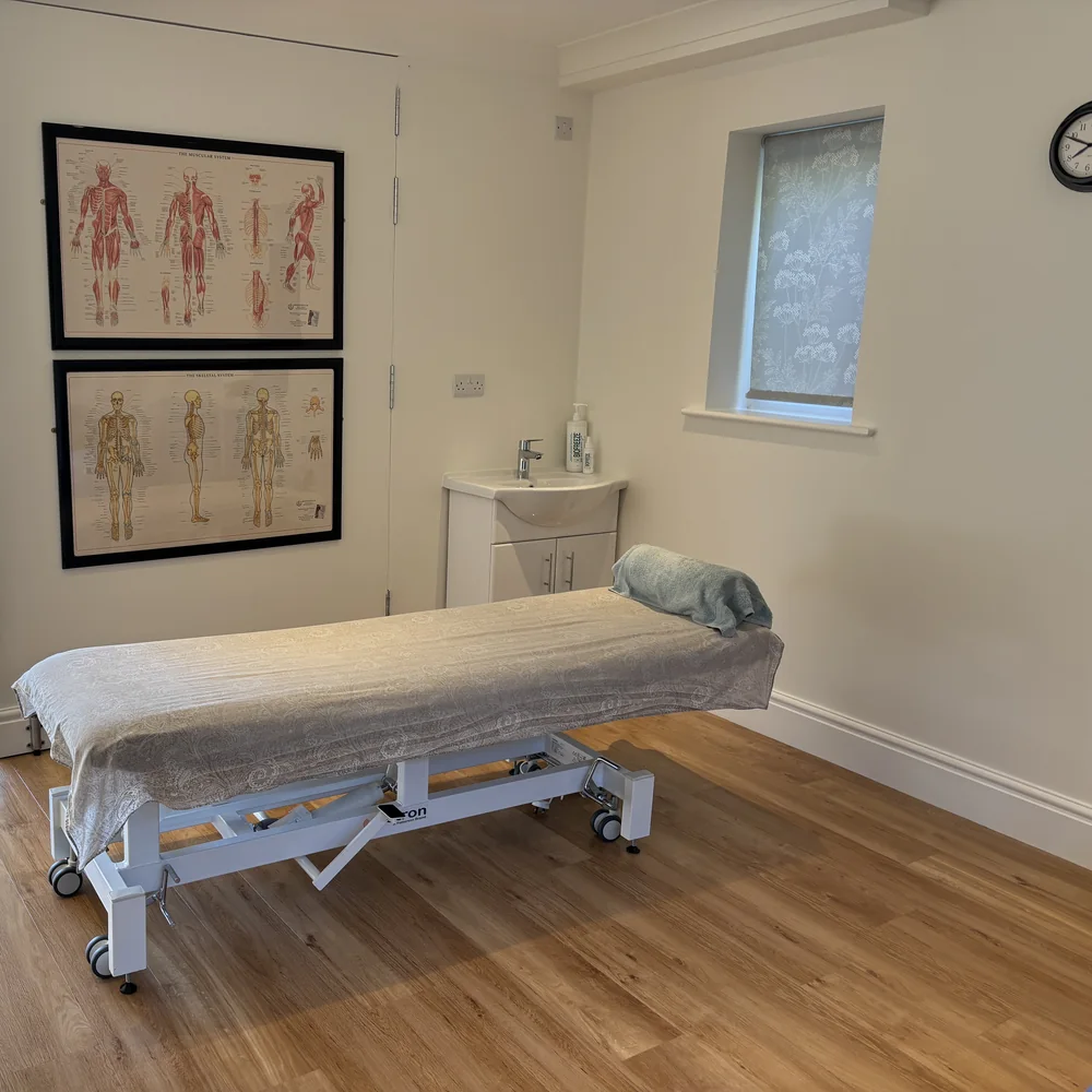 The hydraulic physio couch side on in the treatment corner, the muscular and skeletal charts on the wall behind it and the corner sink beside the door